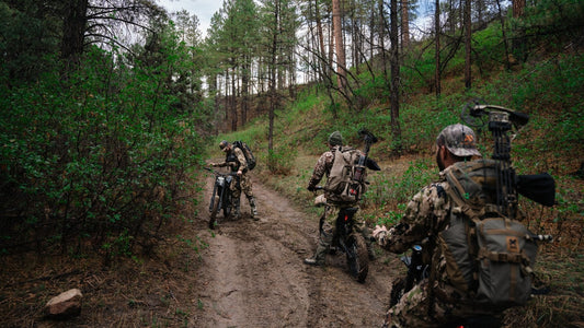 Bowhunters with packs and electric dirt bikes on a muddy, rutted trail through New Mexico pine timber